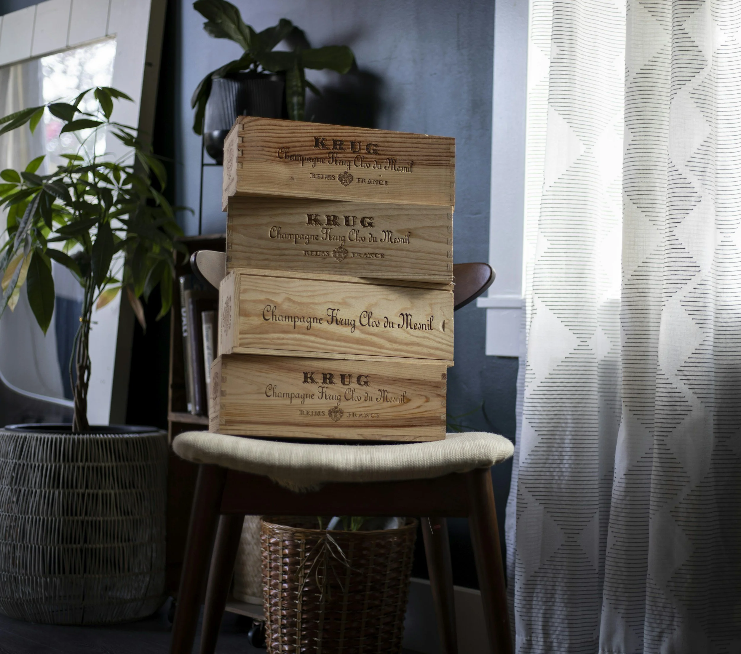 Stacked wooden wine boxes on a chair next to houseplants and a curtain in a cozy interior setting.
