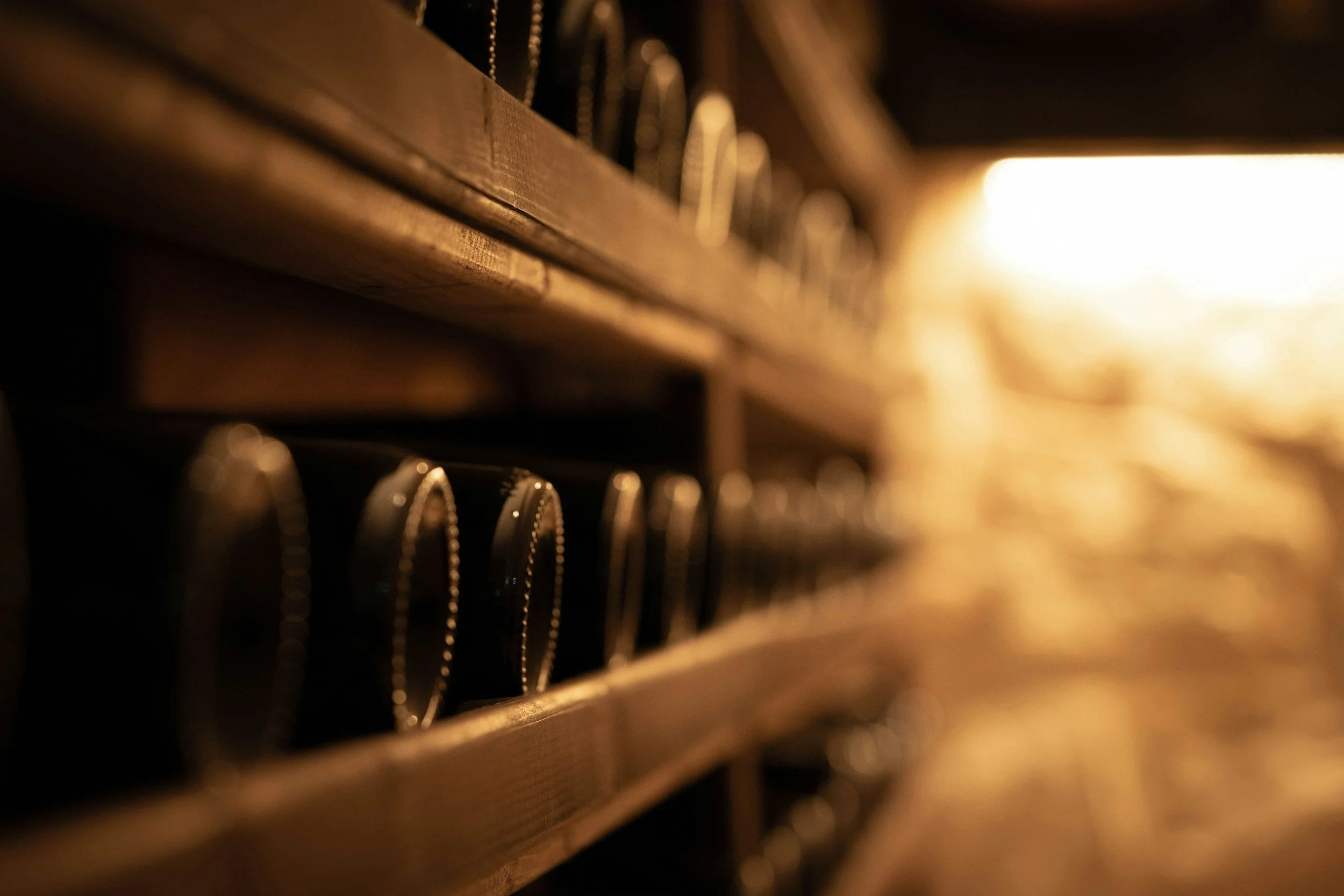 Close-up of wine bottles stored on wooden shelves in a dimly lit wine cellar. Parisian-style wine racks with a warm glow from the light in the background.