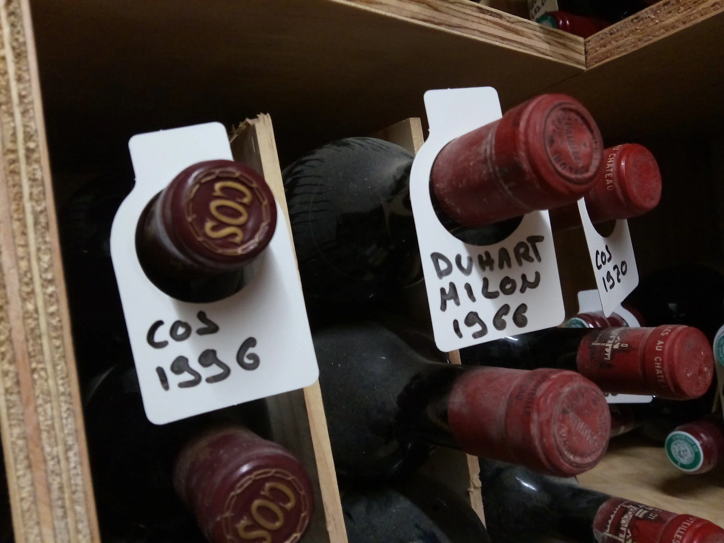 Wine bottles stored in a wooden wine rack with handwritten labels indicating their contents and vintage years, including 'COS 1956' and 'DUHART MILON 1966'.