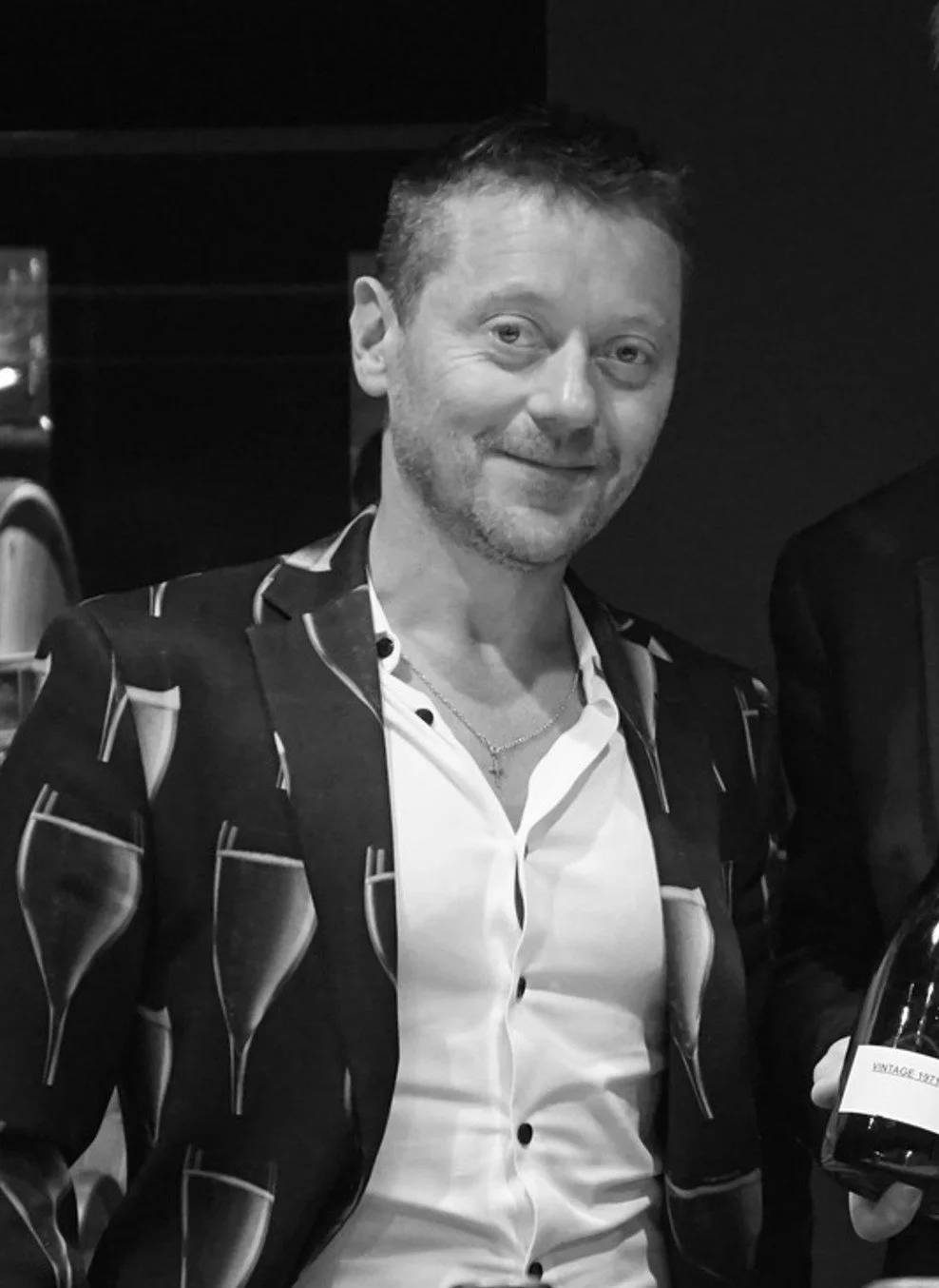 A smiling man with short hair and a trimmed beard, wearing a patterned blazer over a white shirt, holding a wine glass in a dimly lit setting.