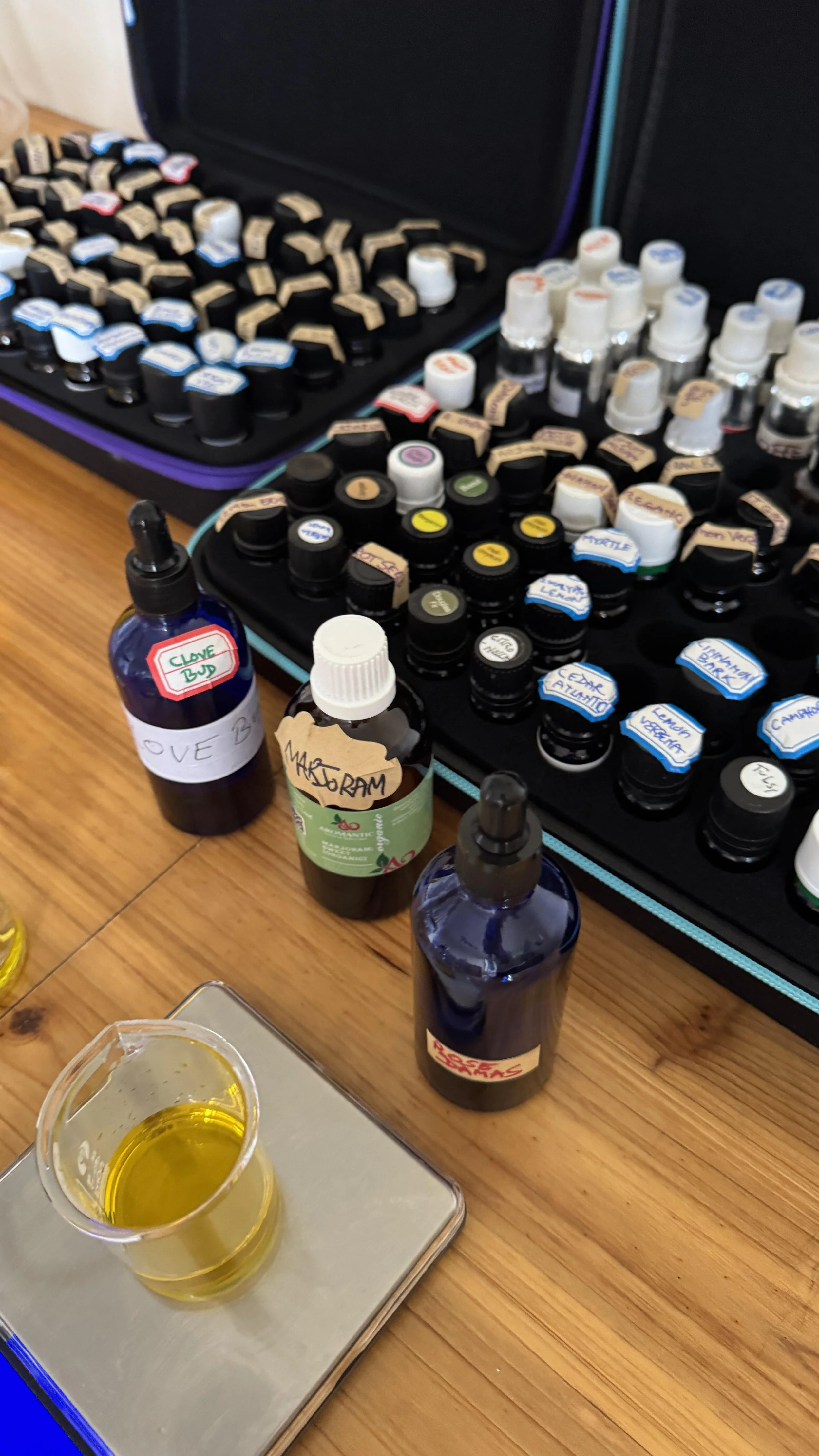 A table with bottles of essential oils, labeled with their scents, in front of open cases filled with additional small bottles, and a small beaker of yellow oil on a scale.