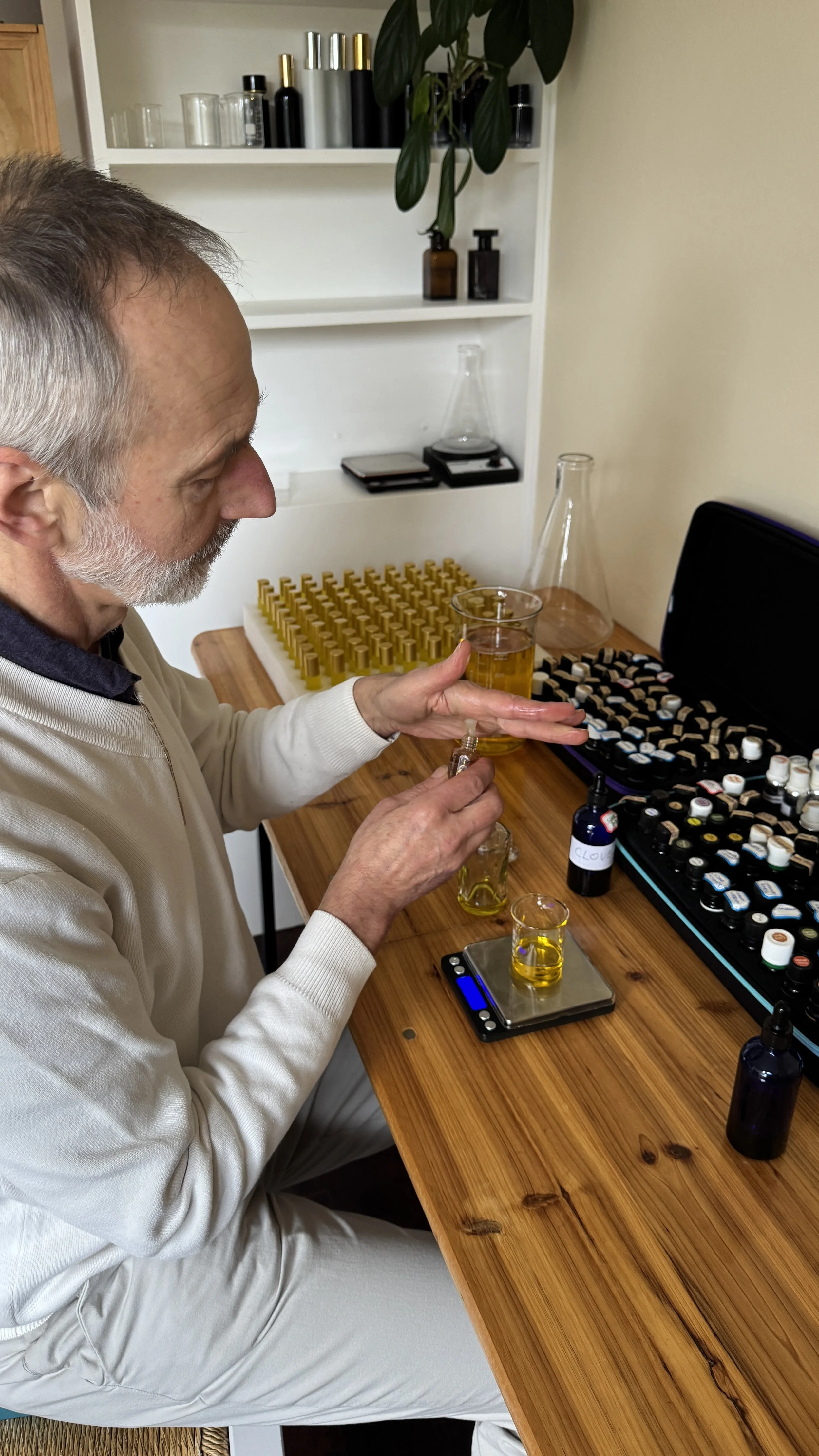 Zeljko Bozic s working with essential oils and small bottles at a wooden table in a laboratory or workshop. There are various glass containers, scales, and a set of small bottles organized on the table and in an open case.