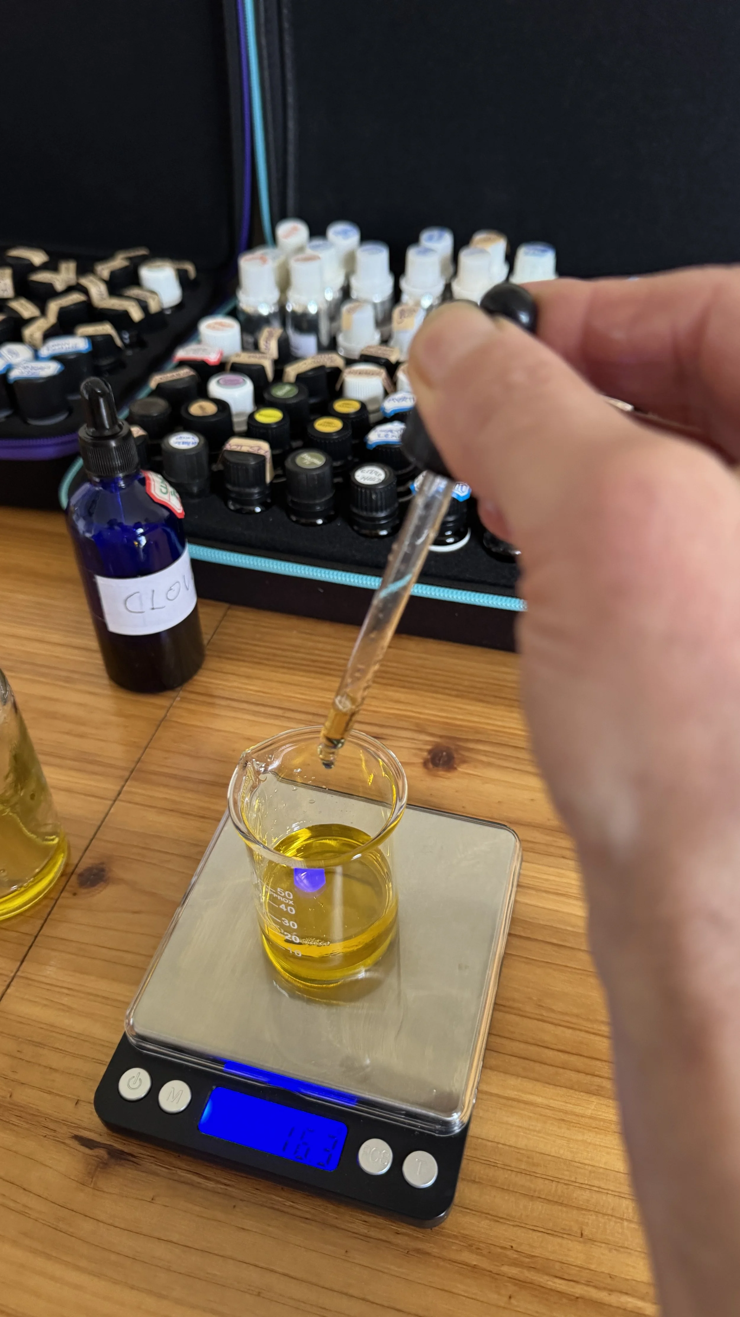 Person using a dropper to transfer yellow liquid into a glass beaker on a digital scale. There are bottles and vials of liquids in the background.