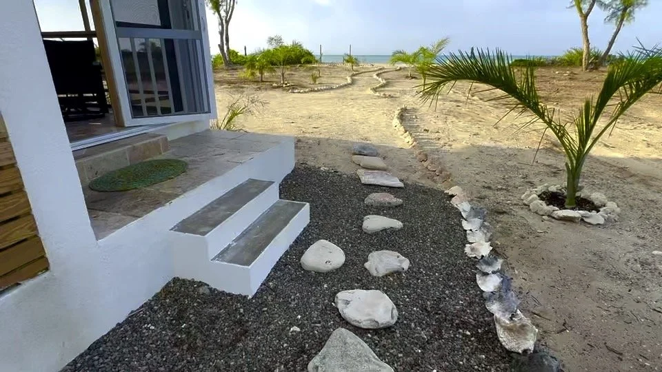Path of flat stones leading from a house step across a small gravel area towards a sandy yard with young palm trees, near the beach.