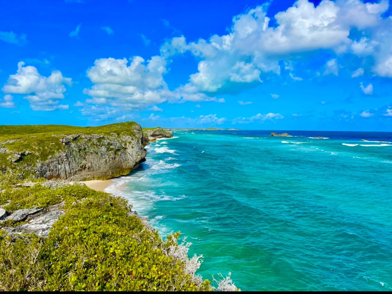 Scenic view of a turquoise ocean with waves crashing against a rocky cliff, green vegetation on the coast, and a partly cloudy sky.