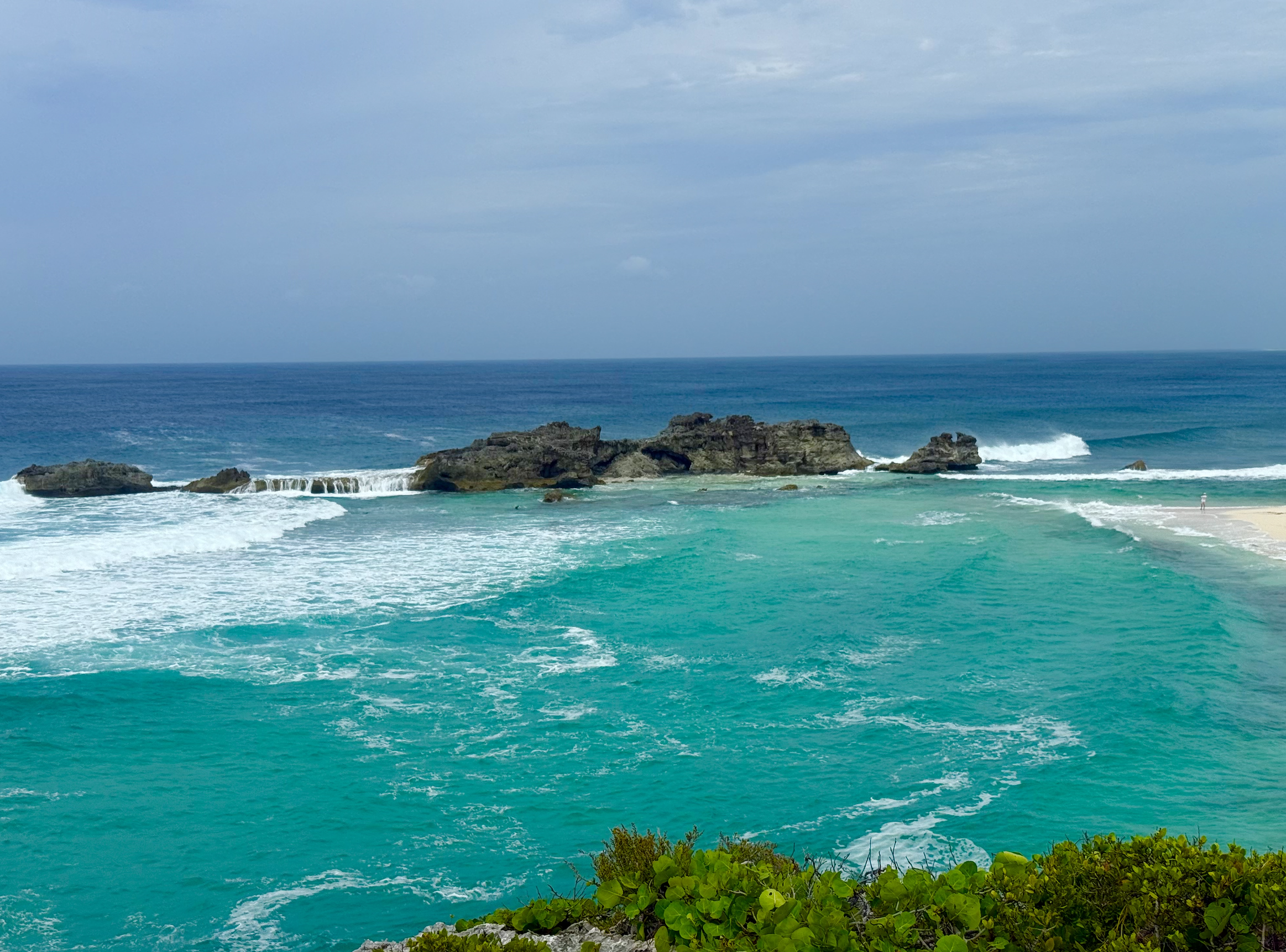 A scenic view of a tropical beach with turquoise waters, rocks in the ocean, and a cloudy sky.