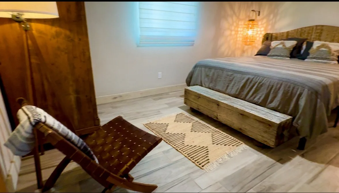 A cozy bedroom with a wooden bed frame, striped bedding, a bench at the foot of the bed, a rug, a wicker lamp on the wall, a window with blinds, and a lounge chair with a blanket.