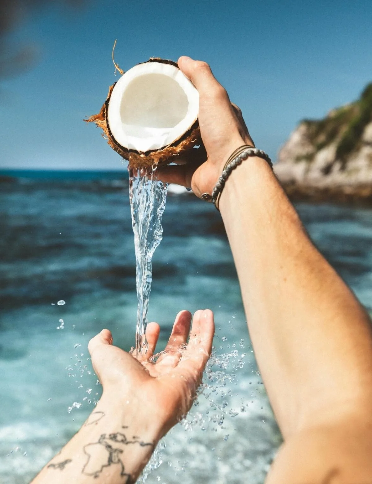 Person holds a half coconut with water pouring out into the ocean on a beach with cliffs in the background