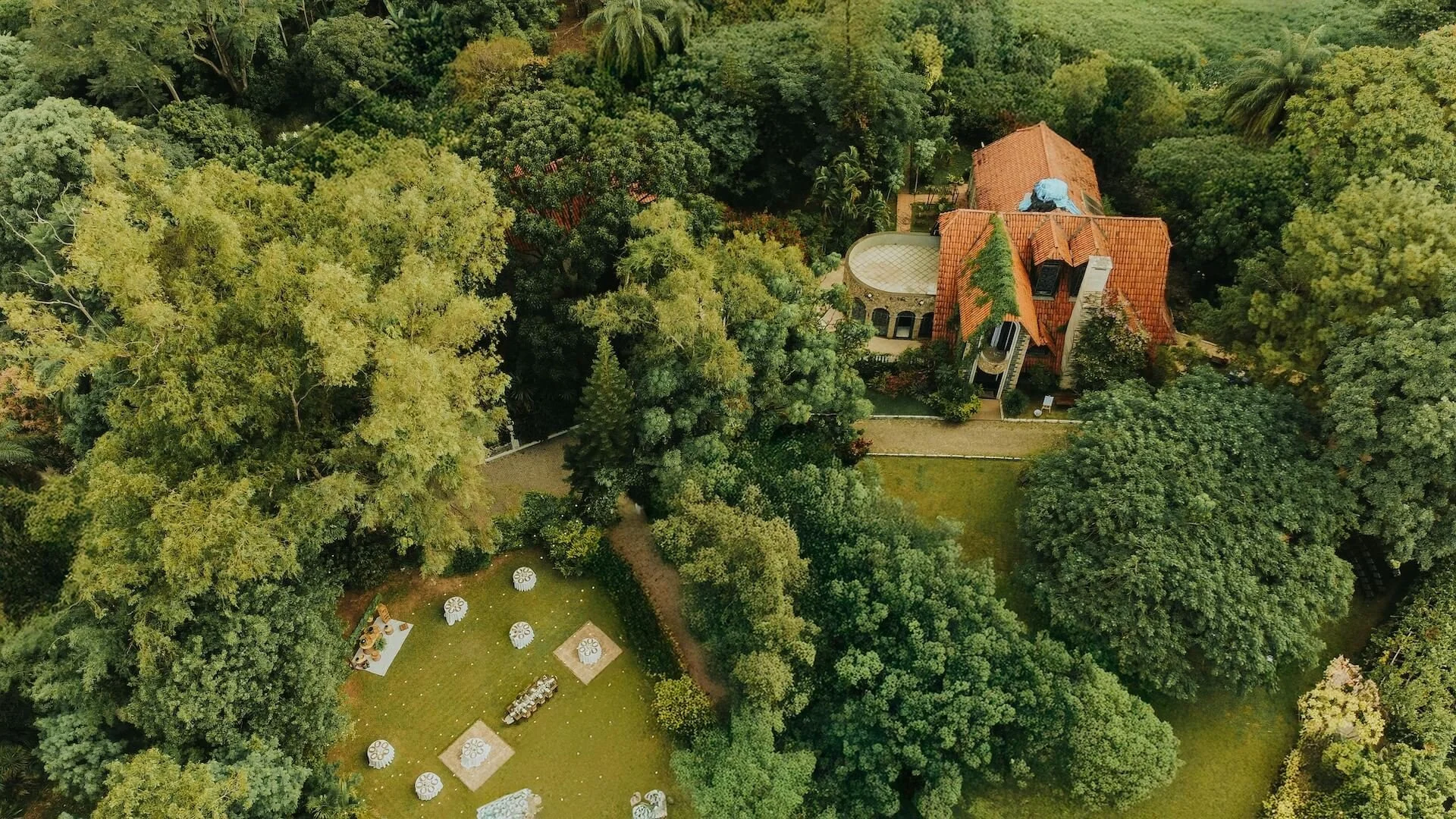 An aerial view of a lush green property with a large house that has a red-tiled roof, surrounded by numerous trees and landscaping, with a backyard area featuring outdoor furniture and umbrellas.