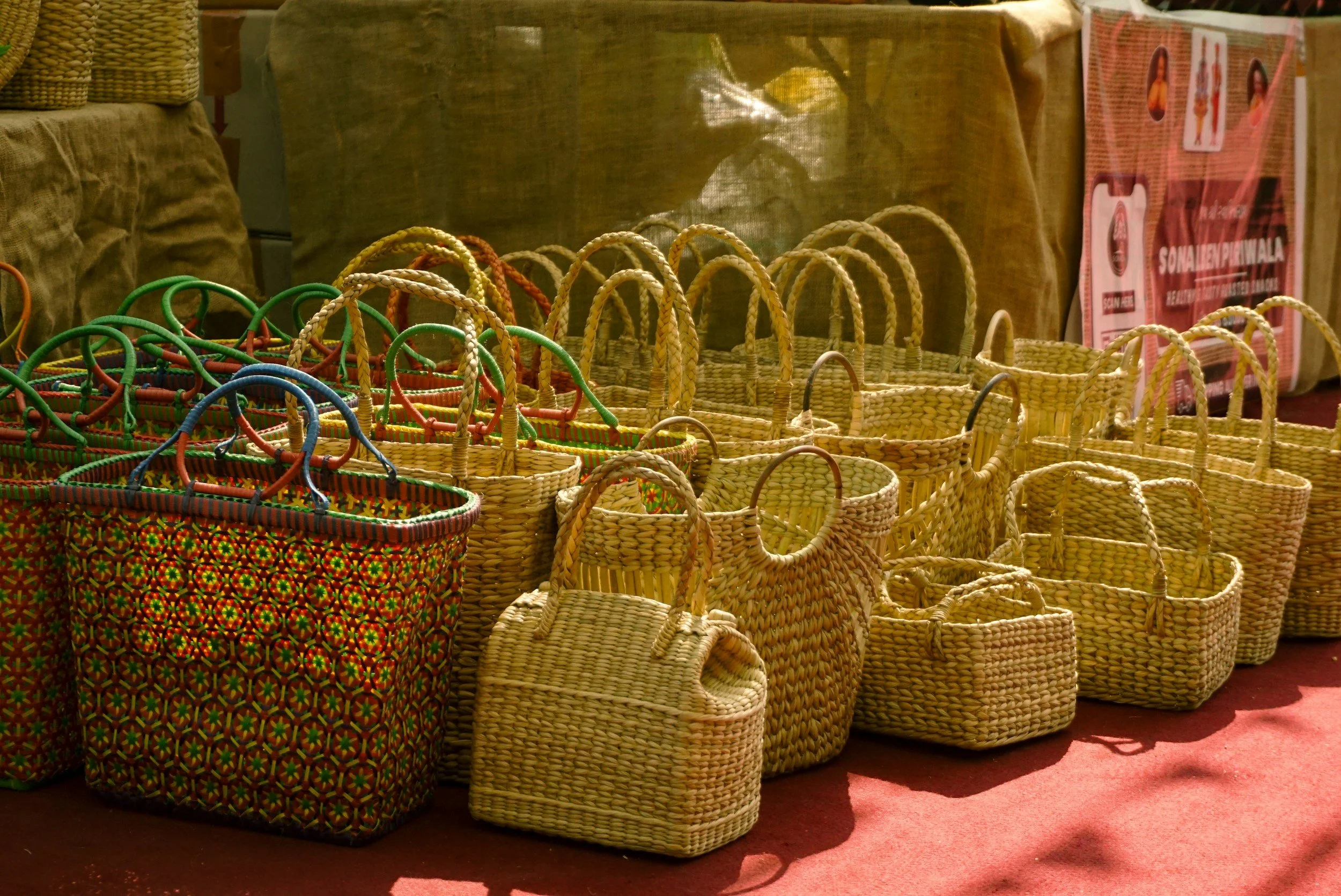 A display of colorful woven baskets of various sizes on a red surface, with a beige cloth background and a poster on the wall.