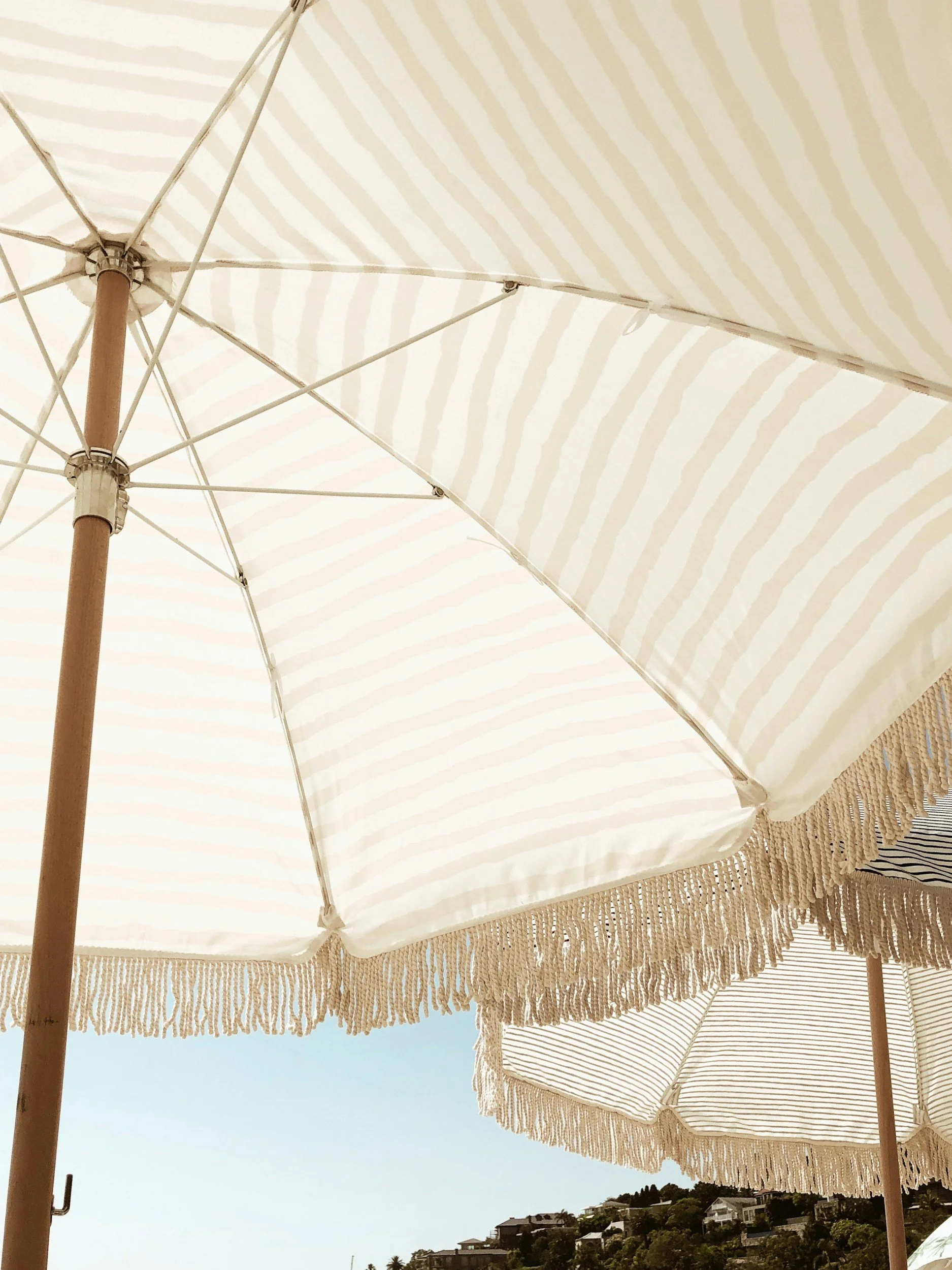 Close-up view of white and beige striped beach umbrellas with fringe edging on a sunny day, with a hillside and houses visible in the background.