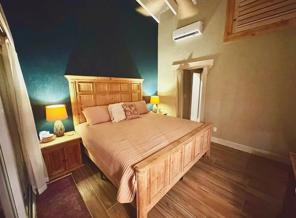 A bedroom with a wooden bed, beige bedding, two bedside lamps, a ceiling fan, an air conditioning unit, and wooden walls and flooring.