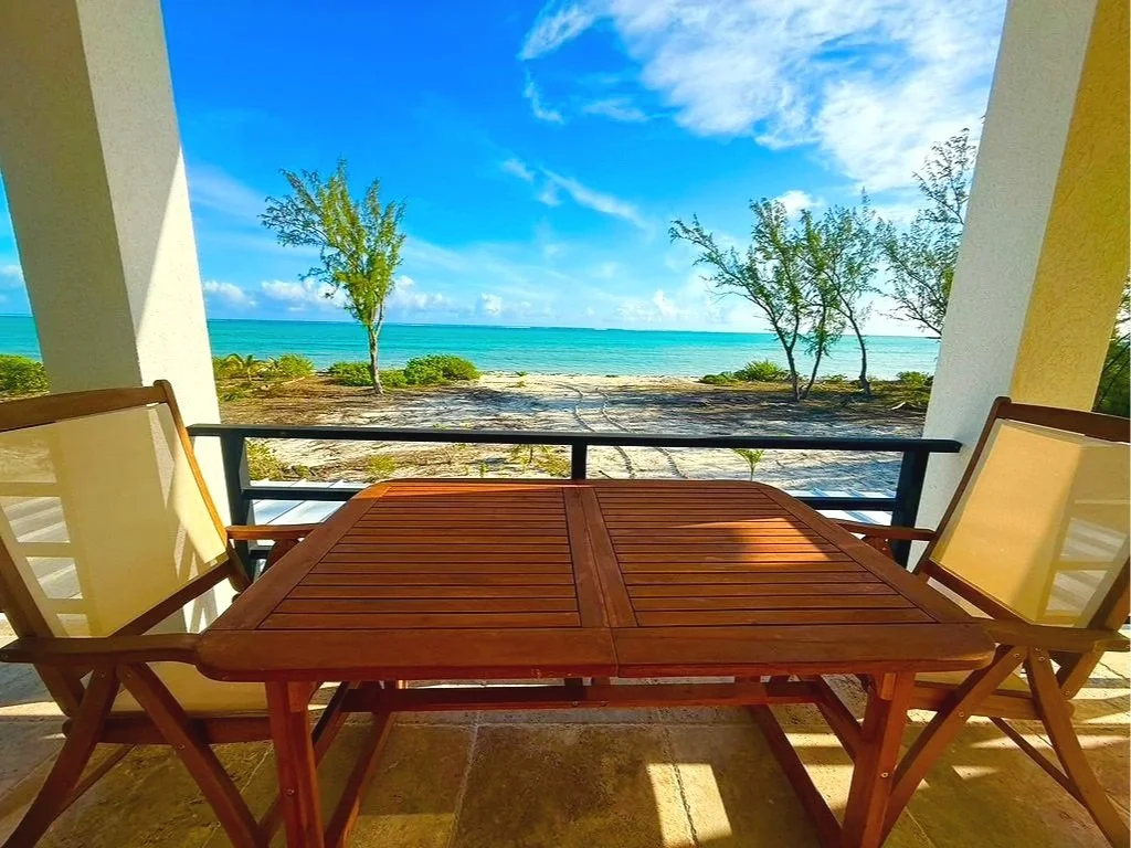 Beach view from a balcony with a wooden table and two chairs, overlooking the ocean with clear blue skies and some trees.