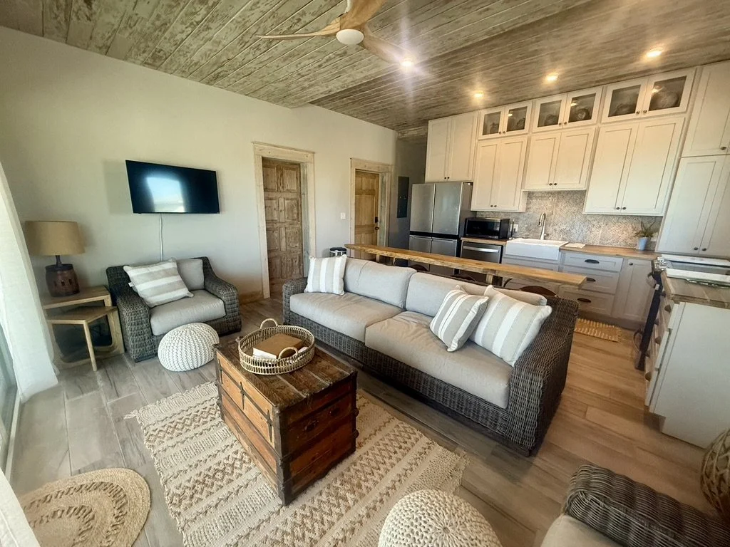 Open-concept living room and kitchen with wicker furniture, a wooden chest coffee table, a wall-mounted TV, and white cabinets with a stainless steel refrigerator.