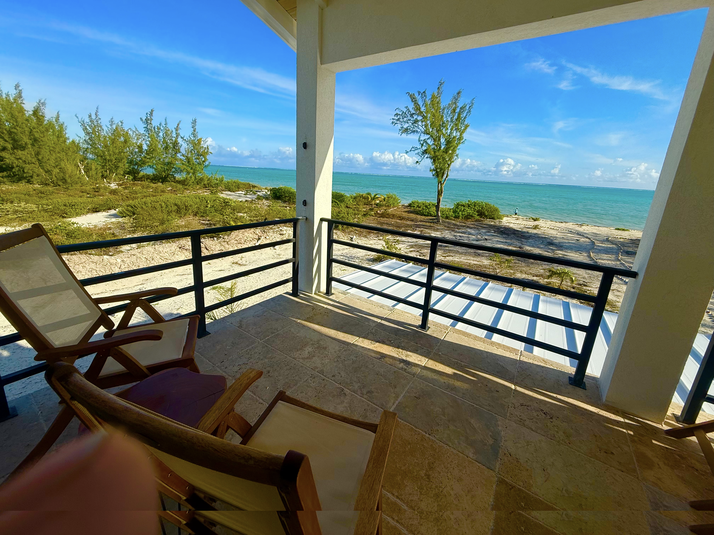 Balcony view overlooking a sandy beach with greenery, a tree, and the ocean under a blue sky with clouds.