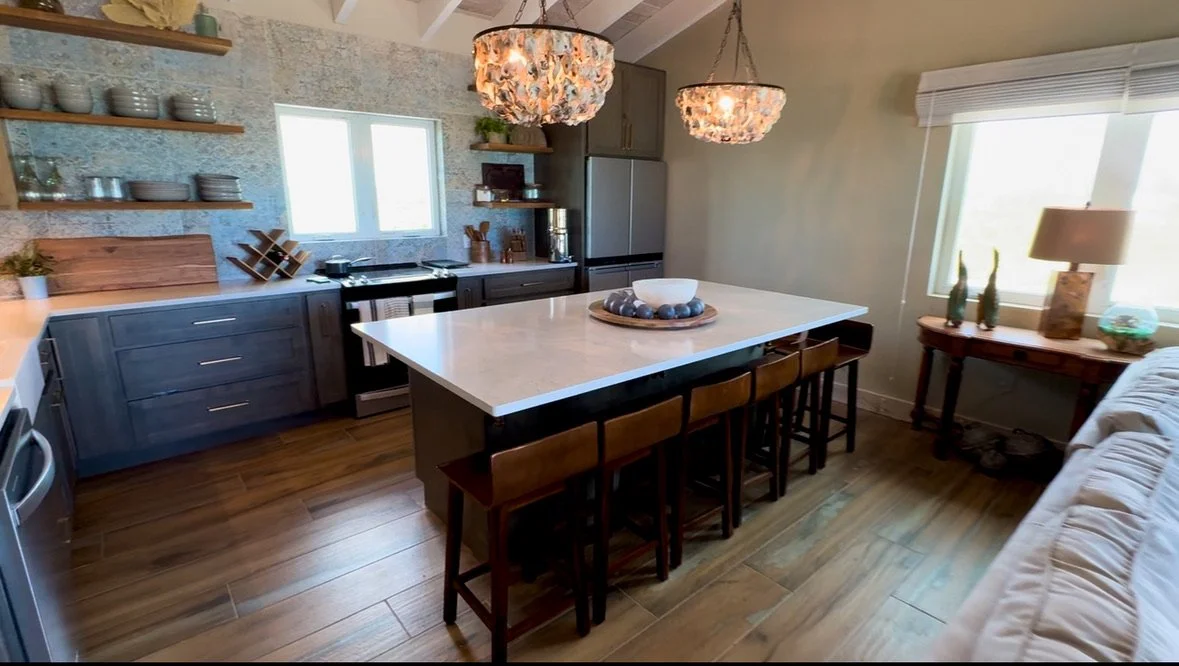 Modern kitchen with wooden flooring, gray cabinets, open wooden shelves with dishes, large window, island with white countertop, four barstools, two pendant lights, side table with two decorative bottles, and a beige sofa.