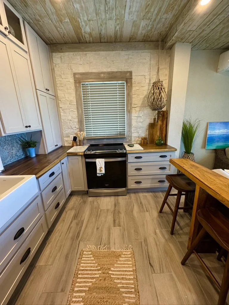 Kitchen with white cabinets, wooden countertops, a black stove, and a window with blinds. There is a small rug on the wooden floor, a potted plant, and a wall with a textured stone finish.