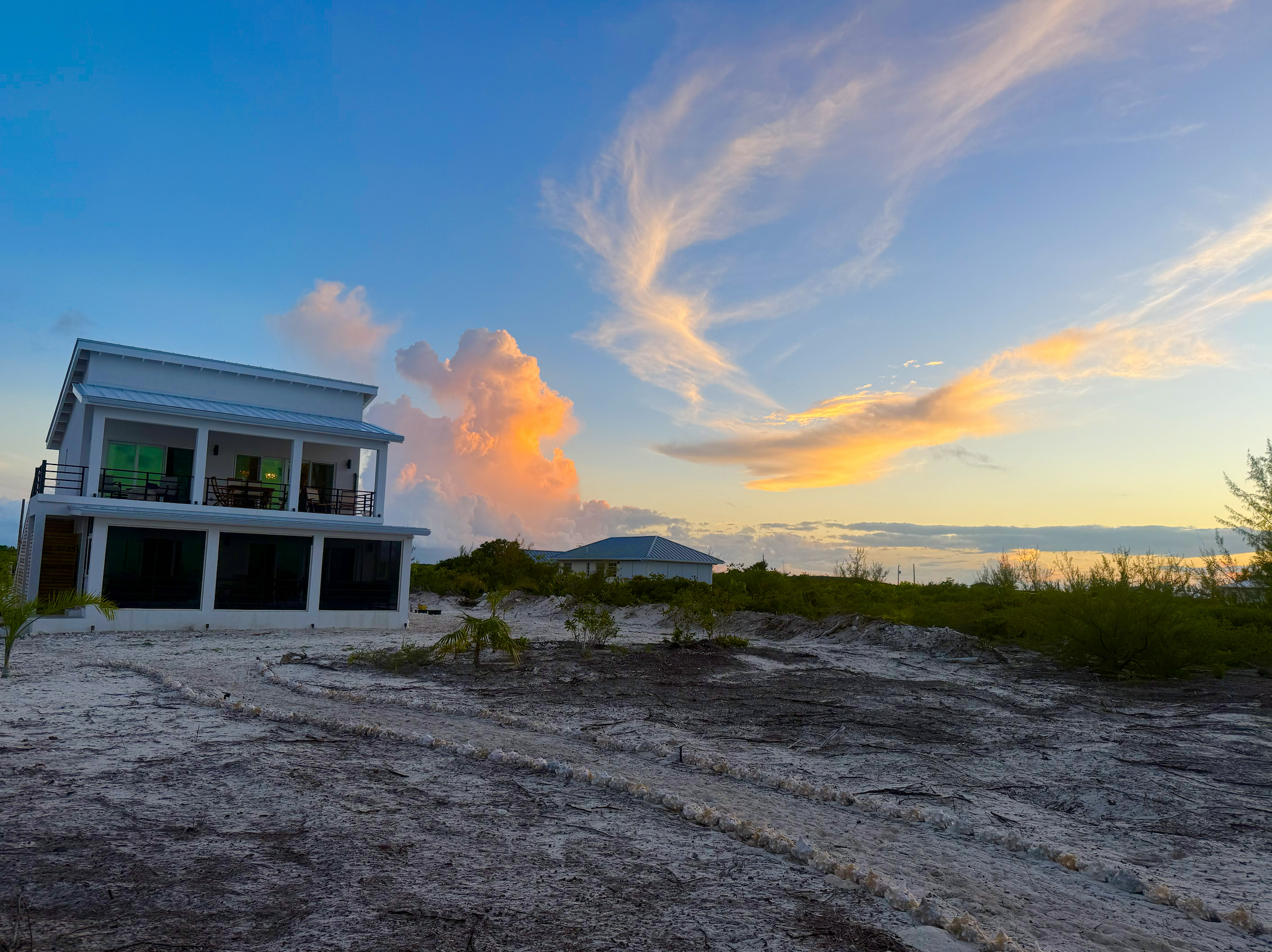 A modern two-story beach house with a white exterior, black railings, and large windows set on sandy terrain with sparse vegetation. The sky is colorful with pink and orange clouds at sunset.