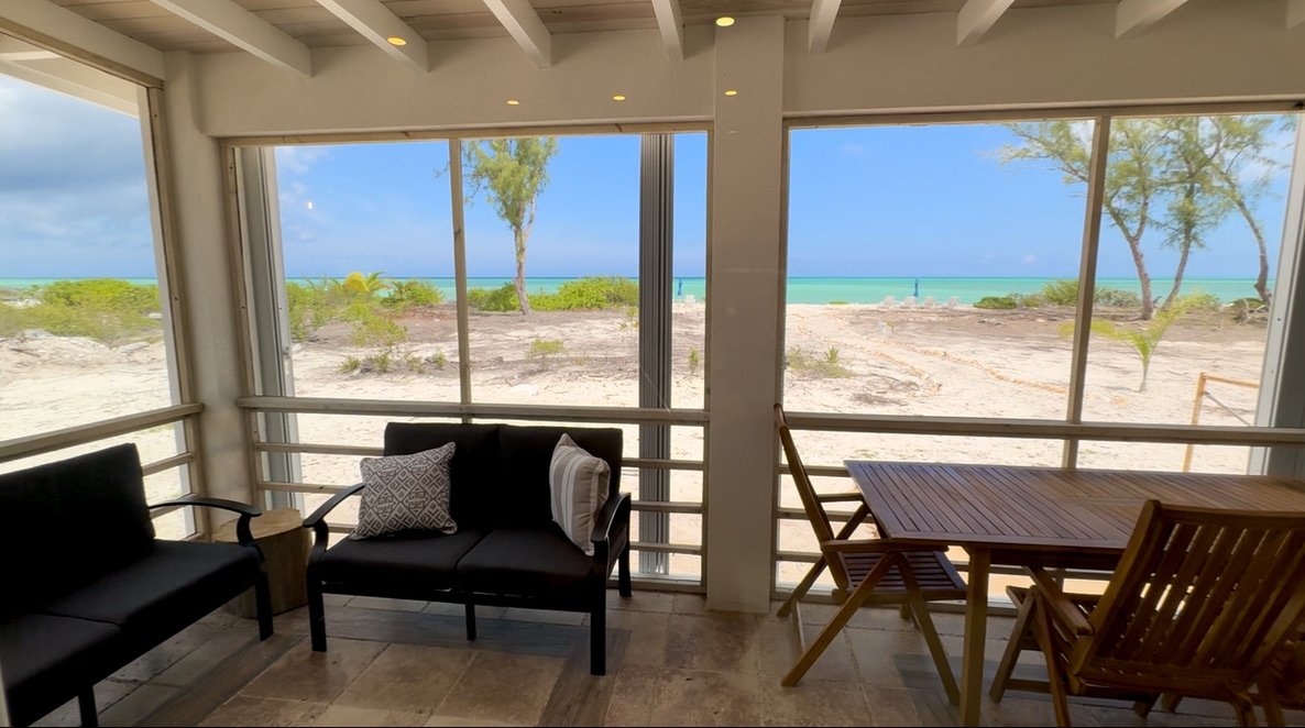 View from a screened porch overlooking a sandy beach with trees, ocean, and blue sky in the background. There are black cushioned chairs with pillows and a wooden table with chairs inside.