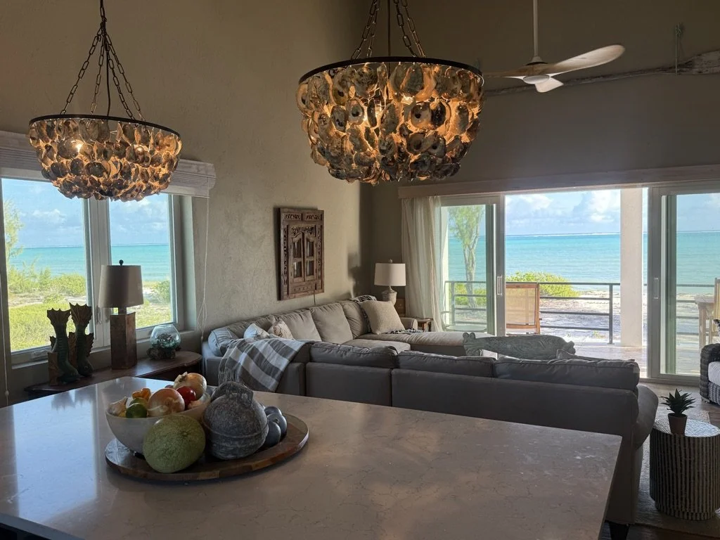Living room with large windows showing a beach view, coastal decor, and two hanging shell chandeliers.