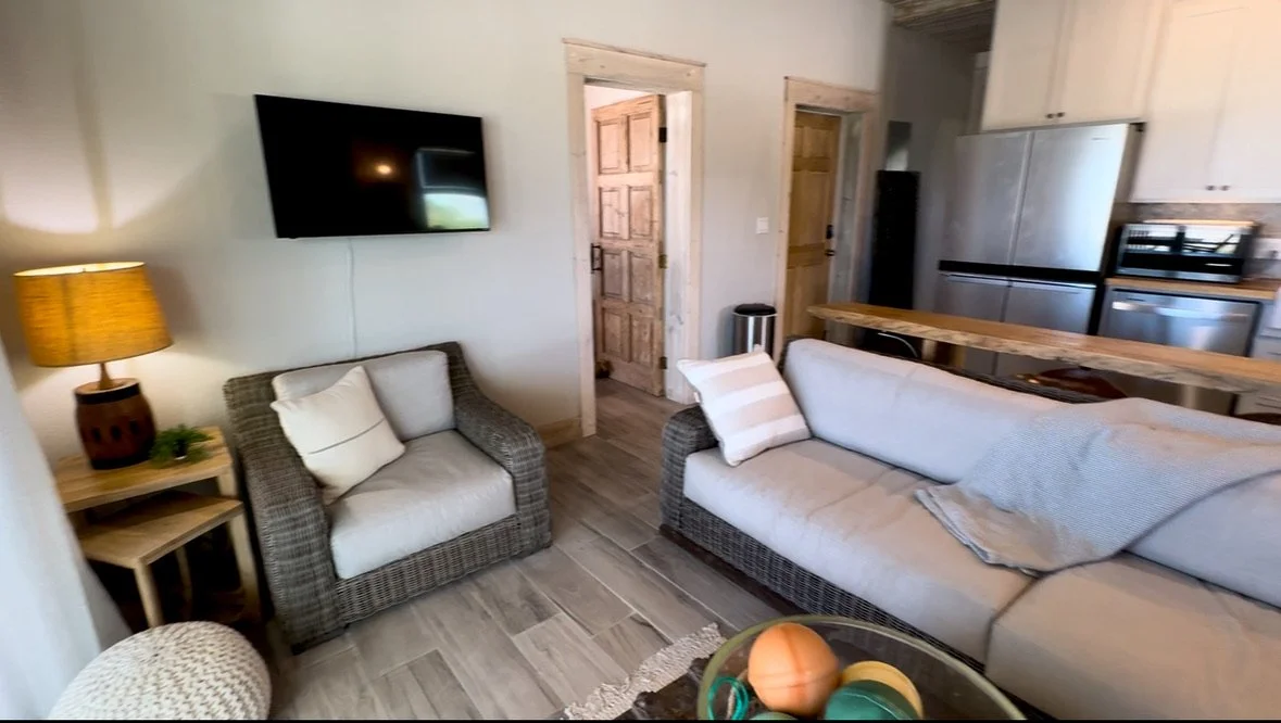 Cozy living room with a wall-mounted TV, a wicker armchair with cushions, a sofa with cushions and a throw blanket, a wooden side table with a lamp and decor, and a kitchen area with a refrigerator and cabinets in the background.