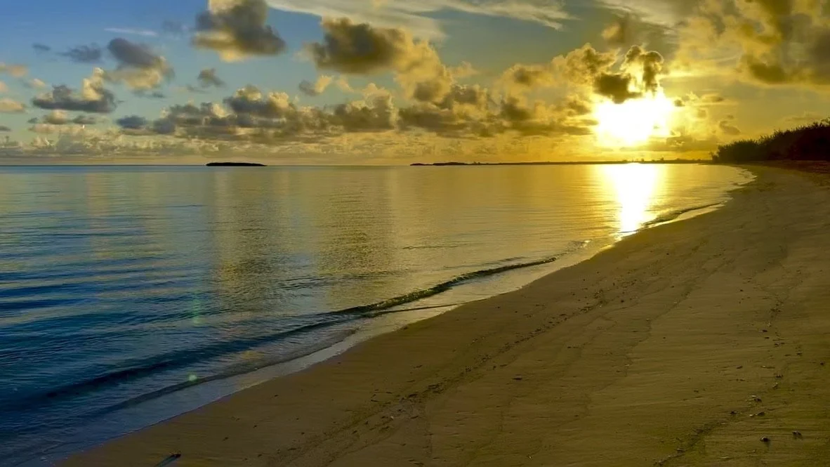 Sunset over a calm beach with a sandy shoreline, gentle waves, scattered clouds, and distant land silhouettes.