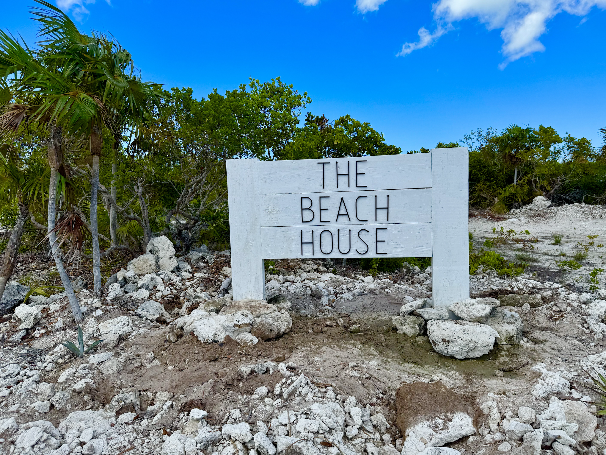 A white wooden sign on rocks reads "The Beach House" with green trees and shrubs, and a bright blue sky with some white clouds in the background.