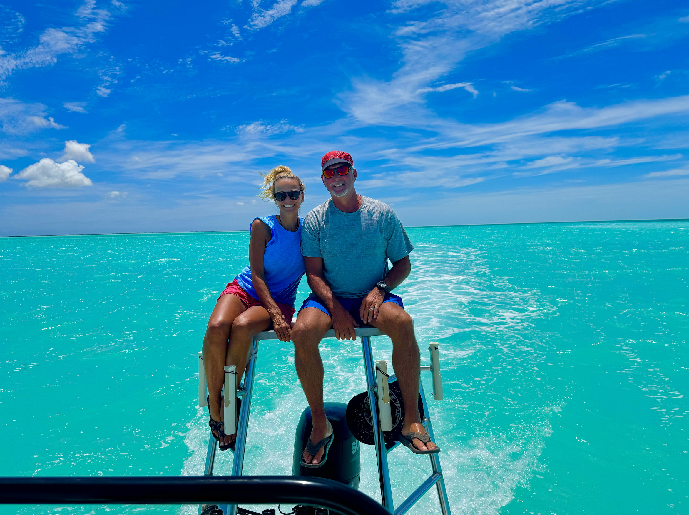 Two people sitting on the edge of a boat, smiling at the camera, with vibrant turquoise water and a partly cloudy blue sky in the background.