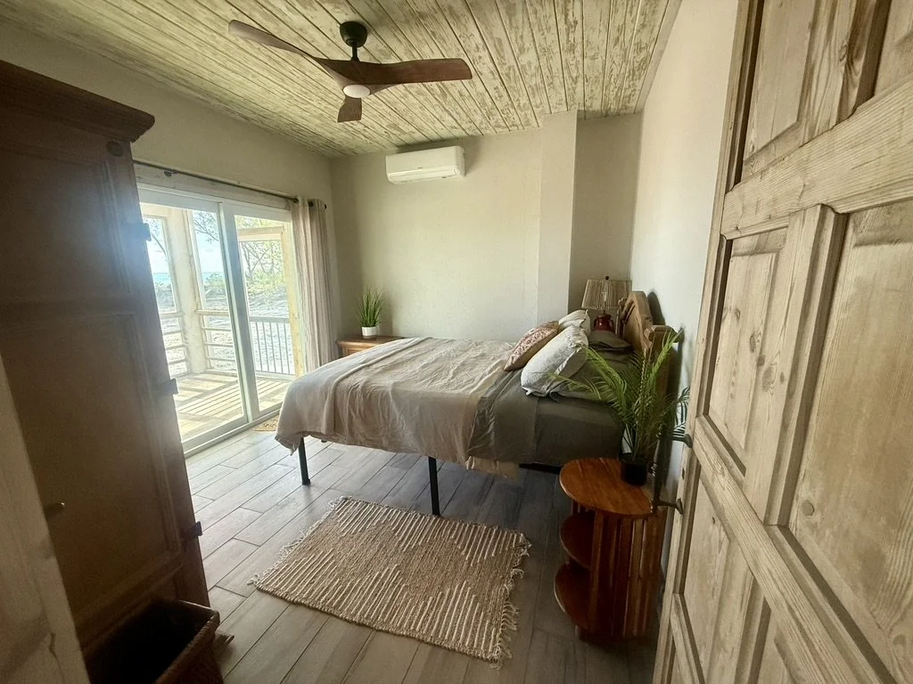 Cozy bedroom with a bed, sliding glass door opening to a balcony, ceiling fan, air conditioner, and wooden furniture.