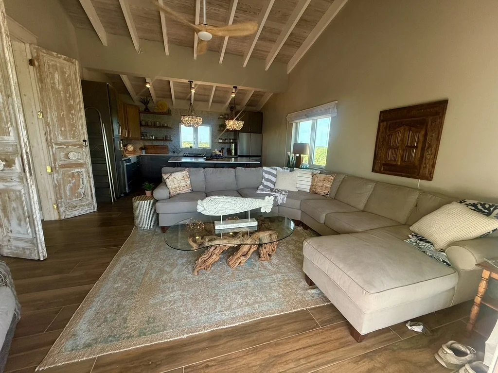 Living room with a large L-shaped beige sectional sofa, glass coffee table with a wooden sculptural base, area rug, and a window with a blind. The background shows a kitchen with pendant lights and a window.