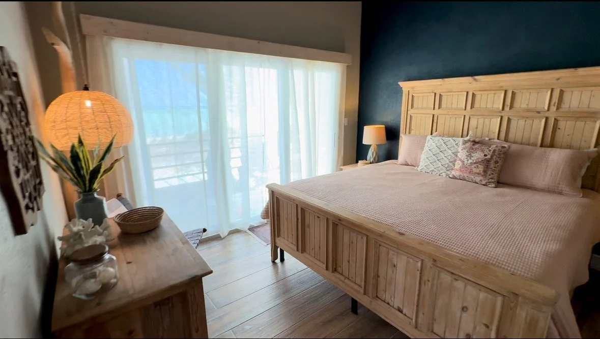 A bedroom with a large wooden bed, pink bedding, and multiple pillows. A nightstand with a lamp and a wooden dresser with decor, next to a sliding glass door with white curtains leading outside.