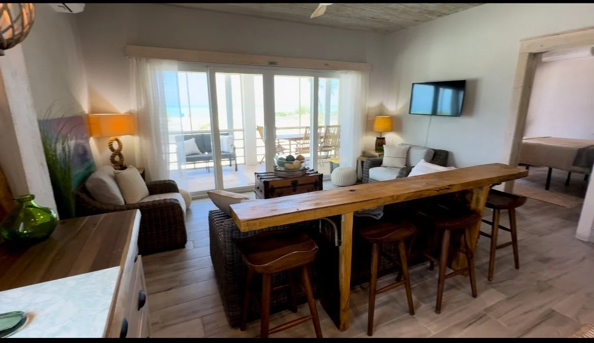 Living room with sliding glass doors leading to a balcony. Contains a wooden countertop with bar stools, a sofa, armchairs, a TV on the wall, and decorative lamps and accessories.