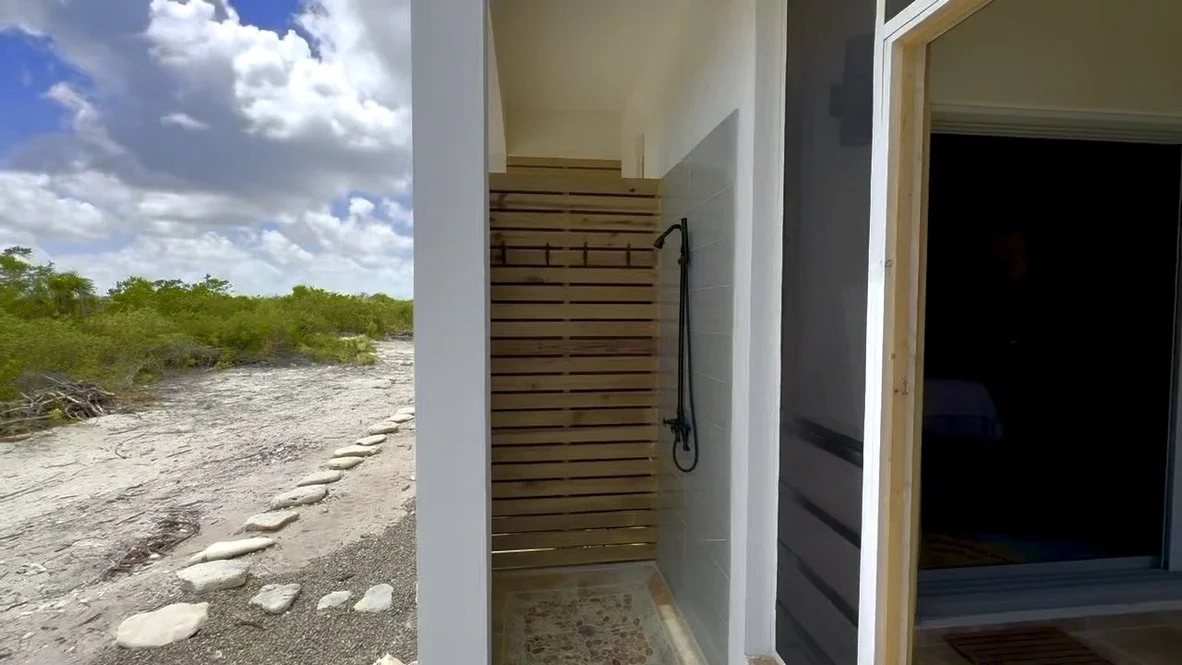 An outdoor shower area with a wooden slatted wall, a black showerhead, and a nearby open door leading inside a house. The landscape has sandy ground with rocks and green bushes, under a partly cloudy sky.