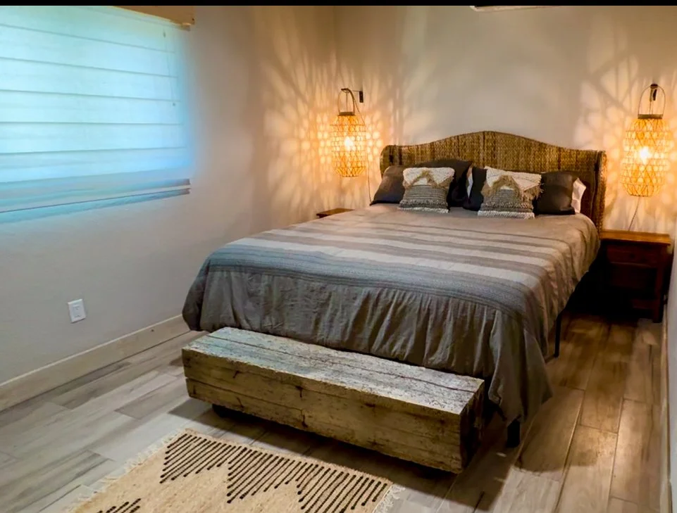 A bedroom with a bed, wooden nightstands, decorative hanging lamps, a wooden chest at the foot of the bed, a window with a white blind, and a patterned rug.