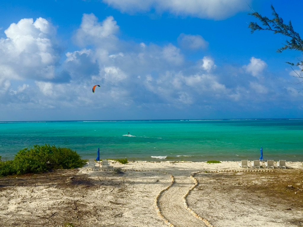 A serene beach scene with a sandy shore, blue umbrellas, lounge chairs, lush greenery, and a bright turquoise ocean with a kite surfer in the distance under a partly cloudy sky.