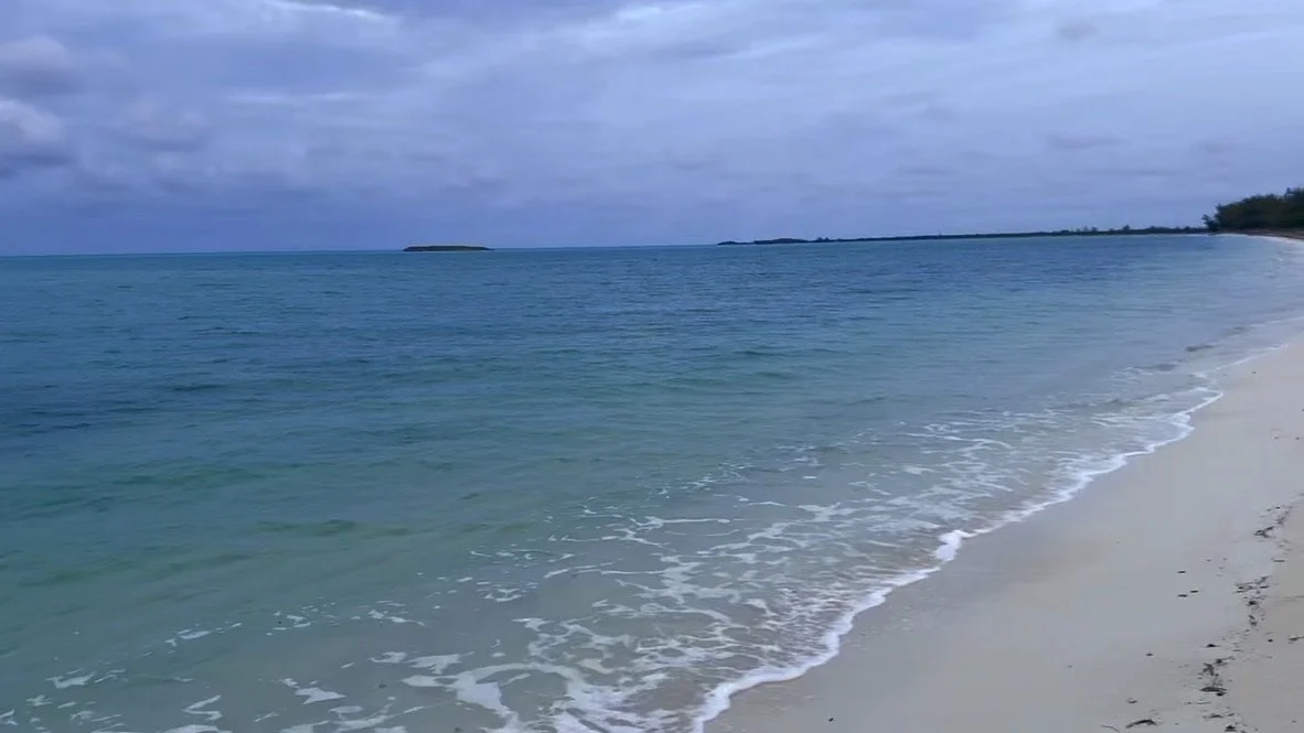 A beach scene with turquoise water, white sand, a cloudy sky, and distant islands on the horizon.