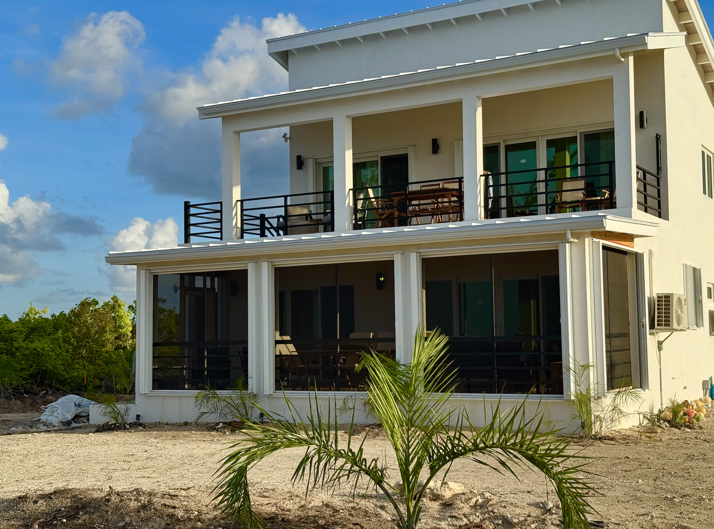 A modern, white, two-story beach house with large screened-in porches on both levels, situated on a sandy lot with a small palm tree in the foreground, under a cloudy sky.