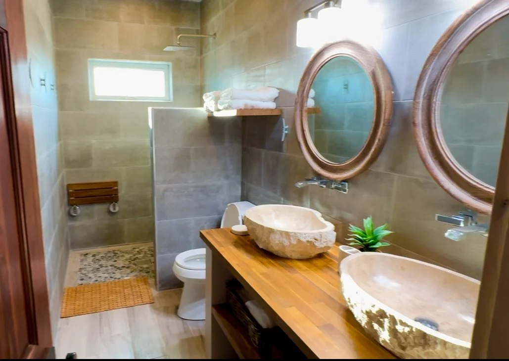 Bathroom with dual stone vessel sinks, round wooden mirrors, tropical plant, bidet, towels, and glass-enclosed shower with a small window.