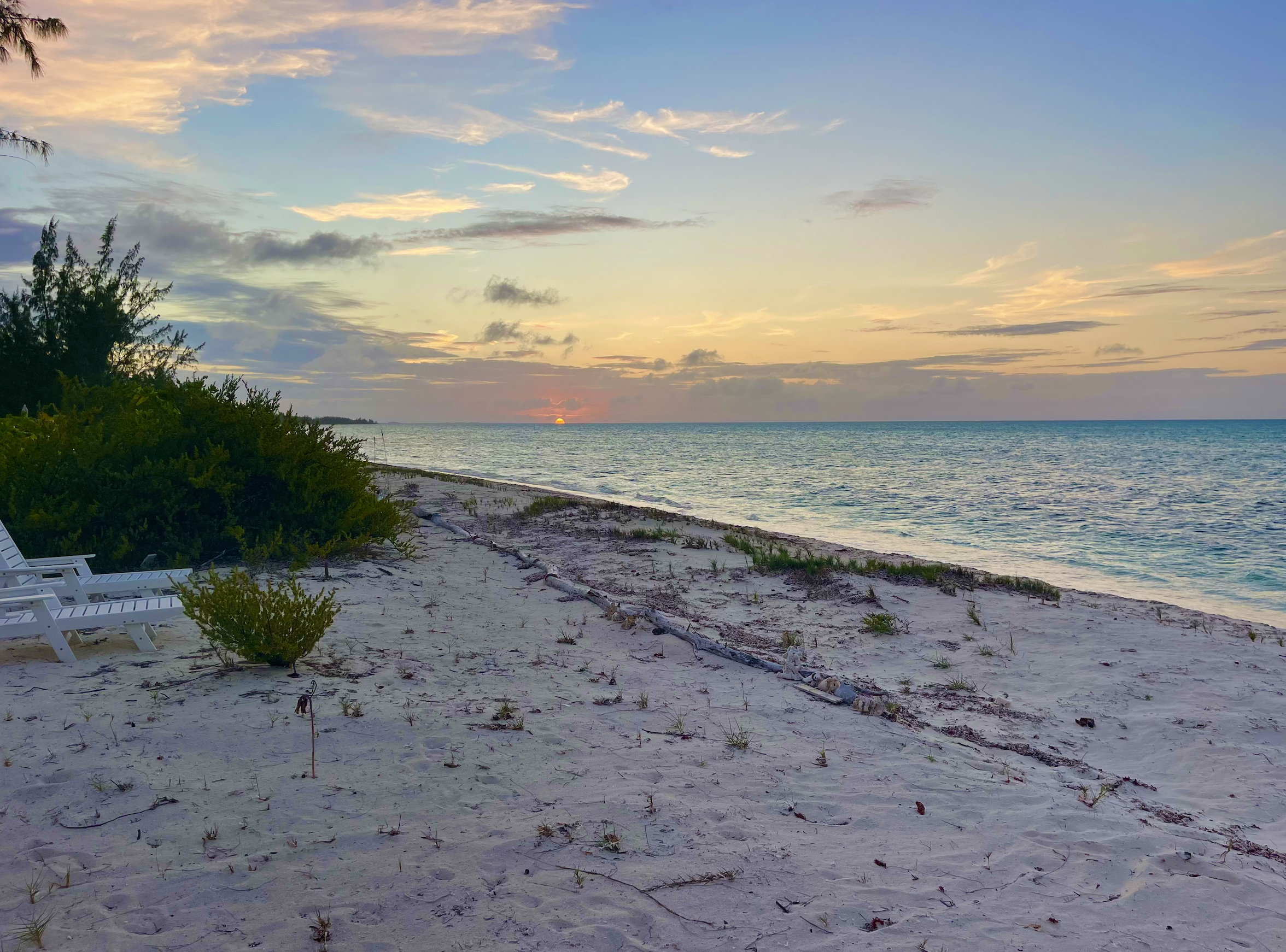 A tranquil beach at sunset with white lounge chairs, green bushes to the left, sandy shore, and calm ocean waves, under a partly cloudy sky.
