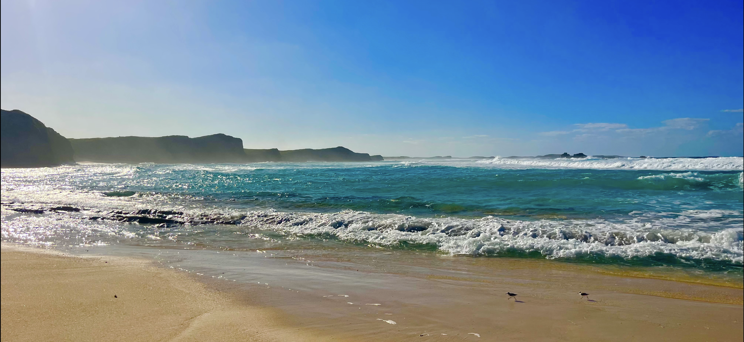 A scenic view of a sandy beach with blue ocean waves breaking near the shore. Cliffs are visible in the background under a bright blue sky.