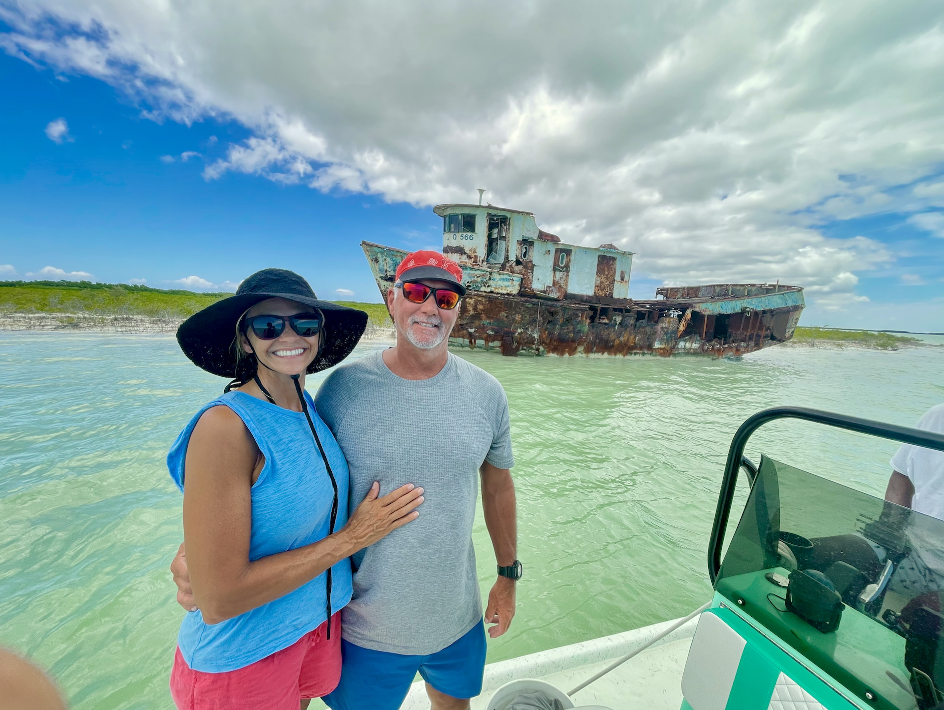Smiling couple standing on a boat, near a rusty, abandoned shipwreck in shallow green water with a cloudy sky overhead.
