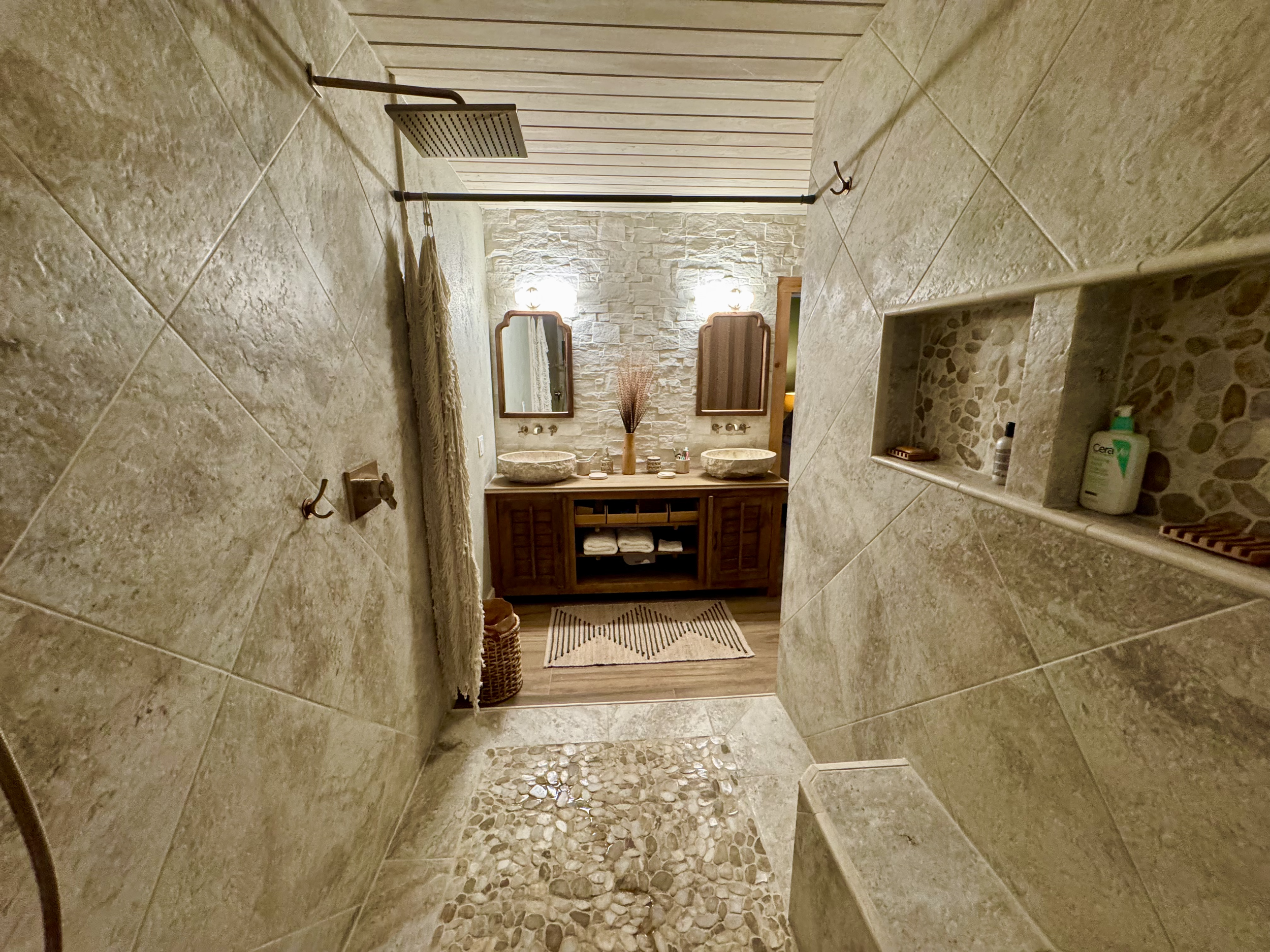 A rustic bathroom with a stone-tiled wall and a pebble floor, featuring a wooden vanity with two vessel sinks, mirrors, and wall-mounted lights.