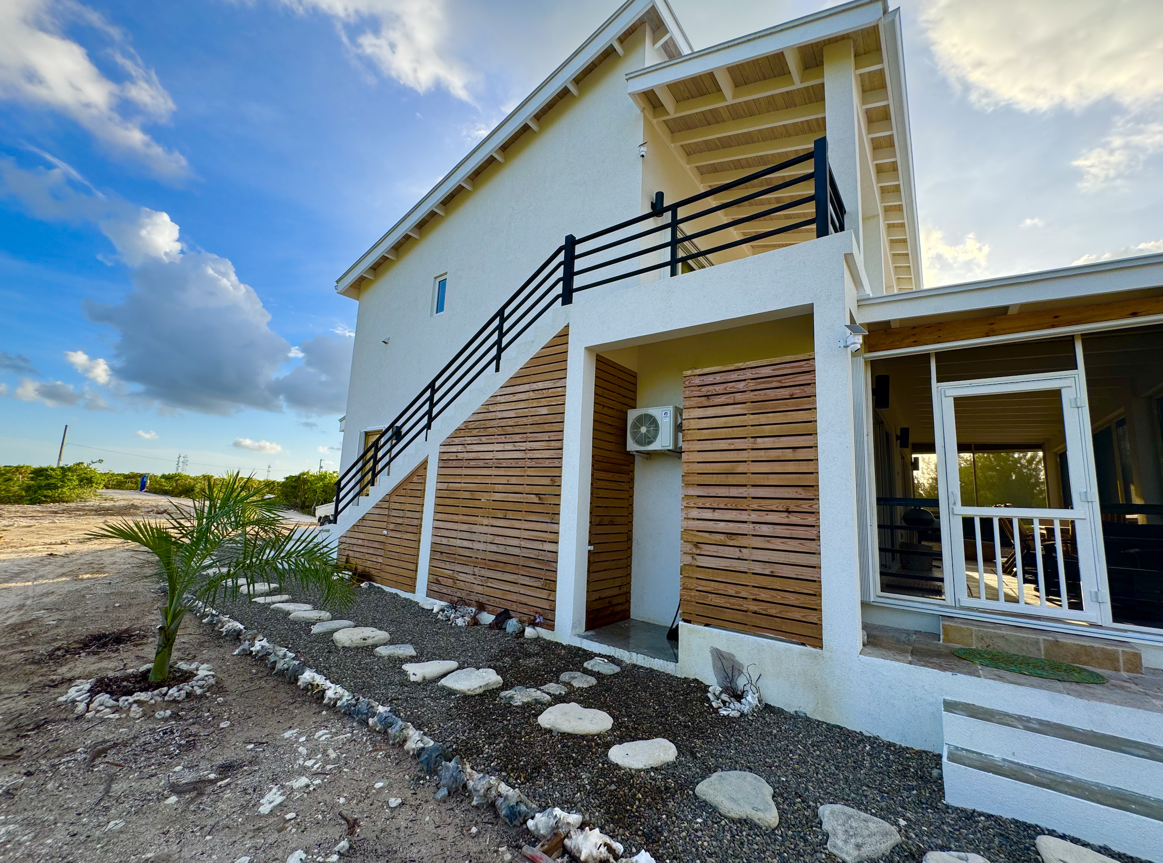 A modern two-story house with white exterior walls and a black railing staircase on the outside, surrounded by a gravel yard with a small palm plant and a stepping stone path, under a partly cloudy sky.