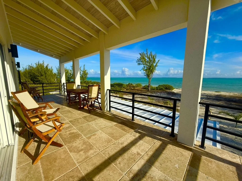 A covered balcony overlooking the beach with several wooden chairs and tables, ocean, trees, and a partly cloudy sky in the background.