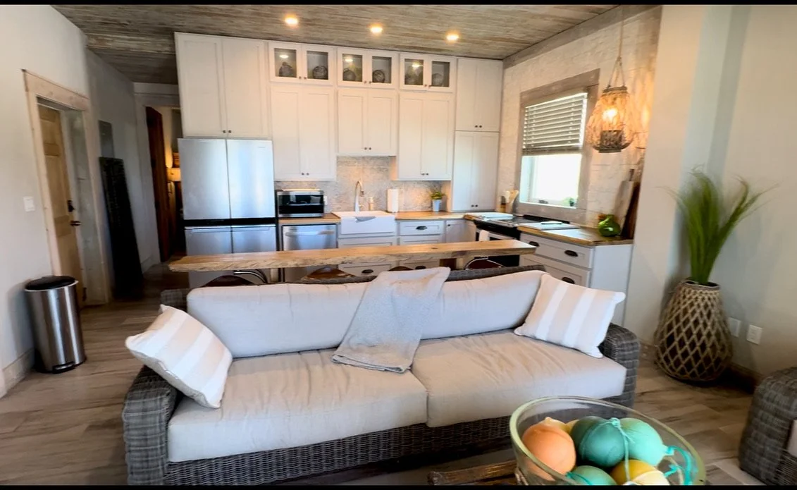 Open-concept kitchen and living room with white cabinets, stainless steel appliances, a wooden kitchen island, a white sofa with striped pillows, potted plant, and decorative hanging lamp.