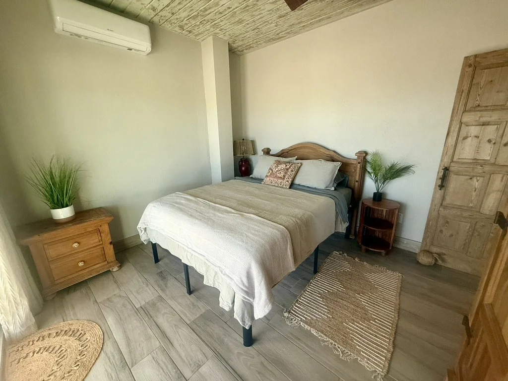 A cozy bedroom with a wooden bed frame, cream bedding, and pillows. There are two small wooden side tables with potted plants, one with a lamp, and a wooden dresser. The room has a wood-paneled ceiling, light-colored walls, and wood flooring. An air 