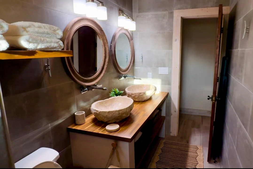 Bathroom with two stone vessel sinks on a wooden vanity, circular mirrors above each sink, wall-mounted light fixtures, folded towels on a shelf, and a partially open door leading to another room.