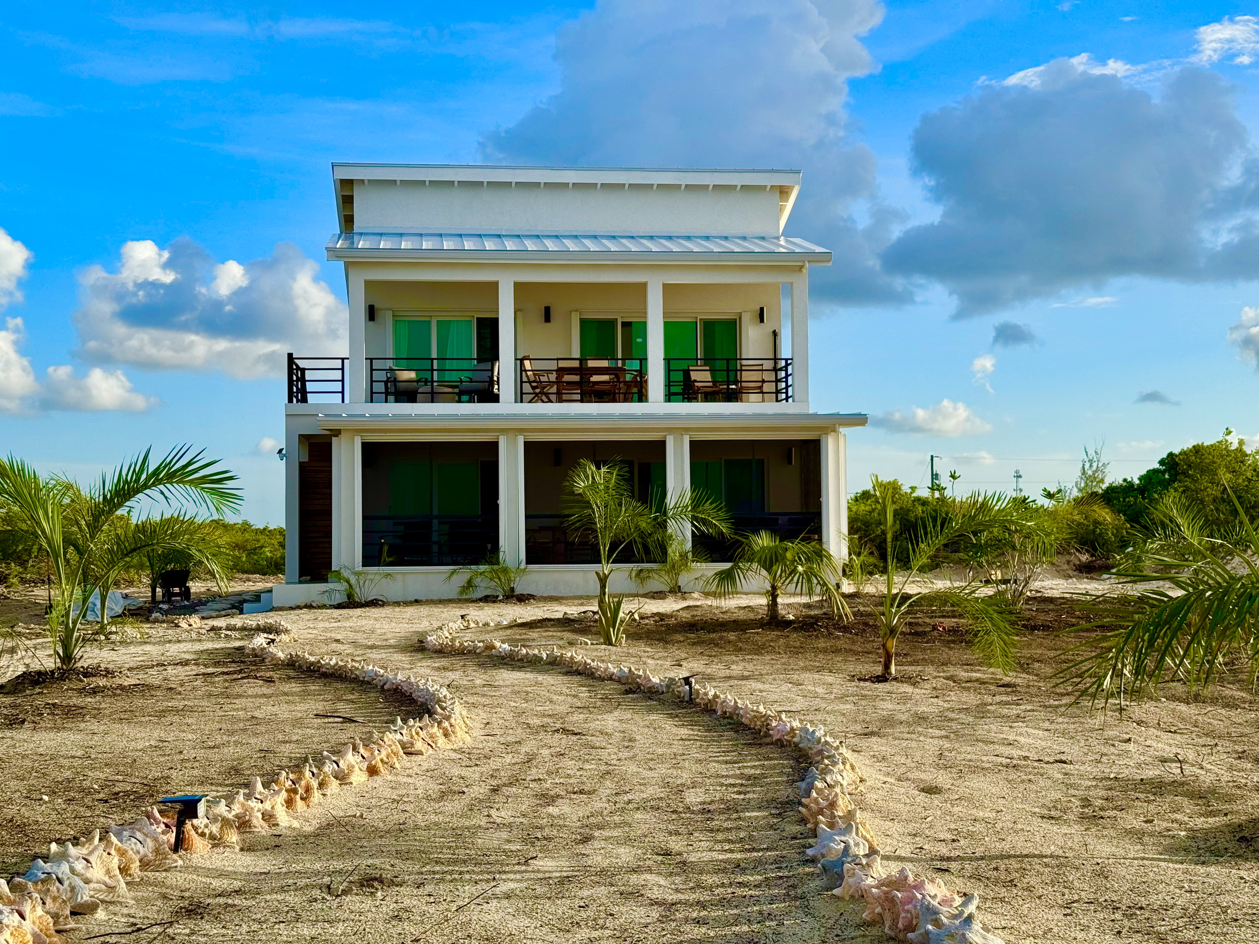 Two-story modern house with large balconies and sliding glass doors, surrounded by small palm trees and shell-lined paths on sandy terrain, under a blue sky with clouds.