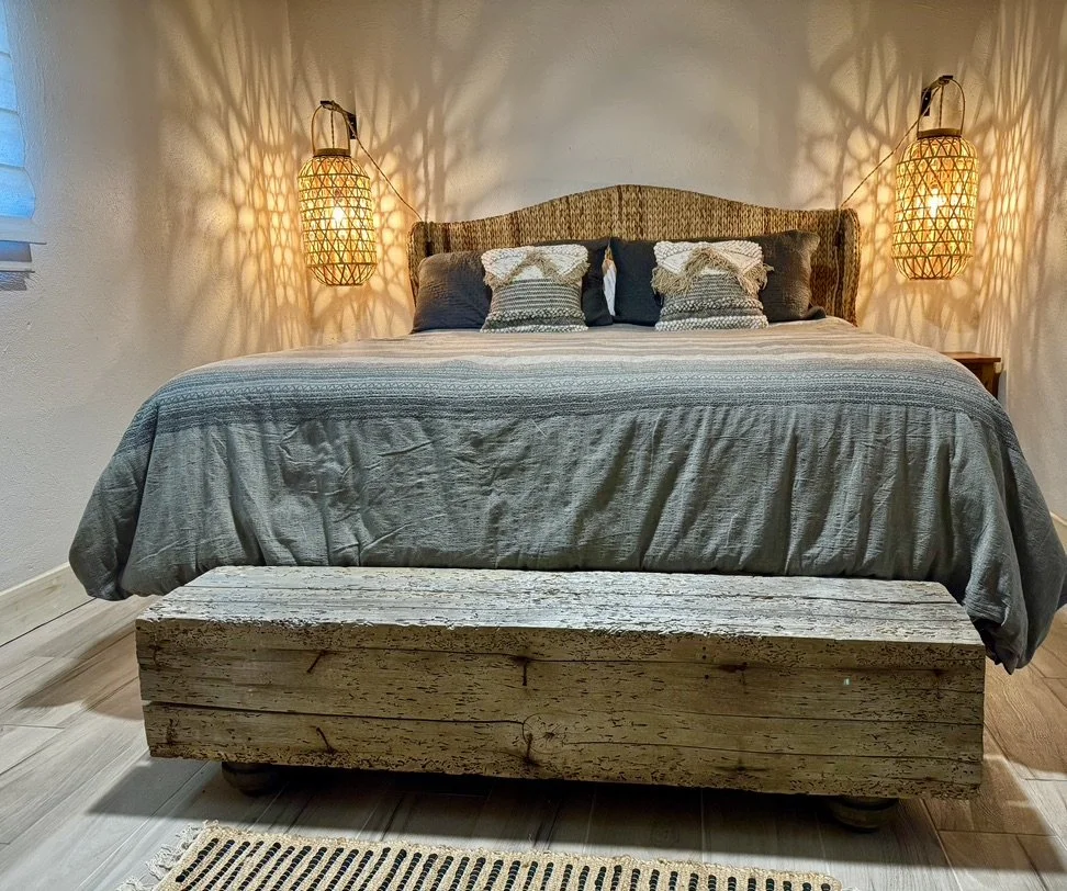 A cozy bedroom with a large bed dressed in gray bedding, accented by decorative pillows. Two woven wall lamps cast warm light and shadows on the white wall behind. At the foot of the bed is a rustic wooden chest. A striped rug is on the light wood fl