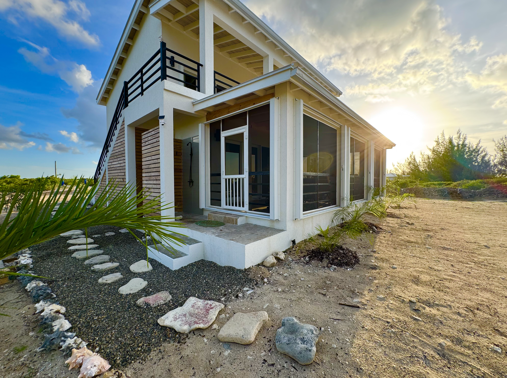 Modern two-story house with large windows and a screened porch, set in a sandy, landscaped yard with rocks and plants, during sunset.